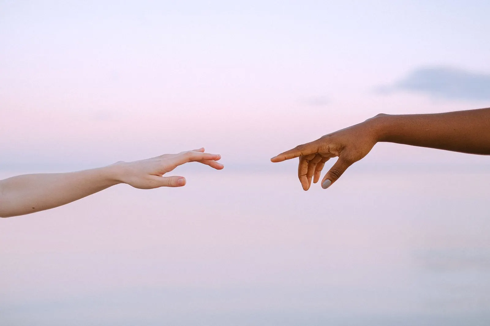Two women's hands reaching towards each other against a soft pastel sky